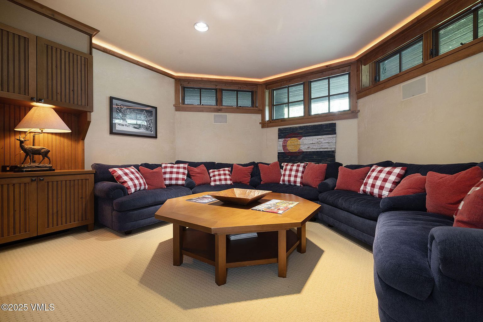 This is an inviting living room featuring a large, comfortable sectional sofa adorned with red and plaid pillows. A unique, multi-sided wooden coffee table sits in the center of the room, complemented by built-in wooden cabinetry and warm lighting. The room exudes a cozy and rustic charm, perfect for relaxation and gatherings.