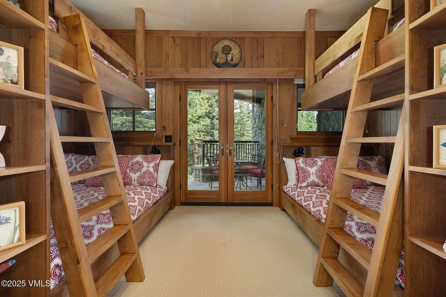 This is an interior shot of a bedroom featuring built-in bunk beds on either side of a set of glass doors leading to a balcony. The room has a rustic, cabin-like feel with wood paneling and matching wooden ladders leading to the upper bunks. The beds are adorned with red and white patterned bedding, and the room is well-lit, creating a cozy and inviting atmosphere.