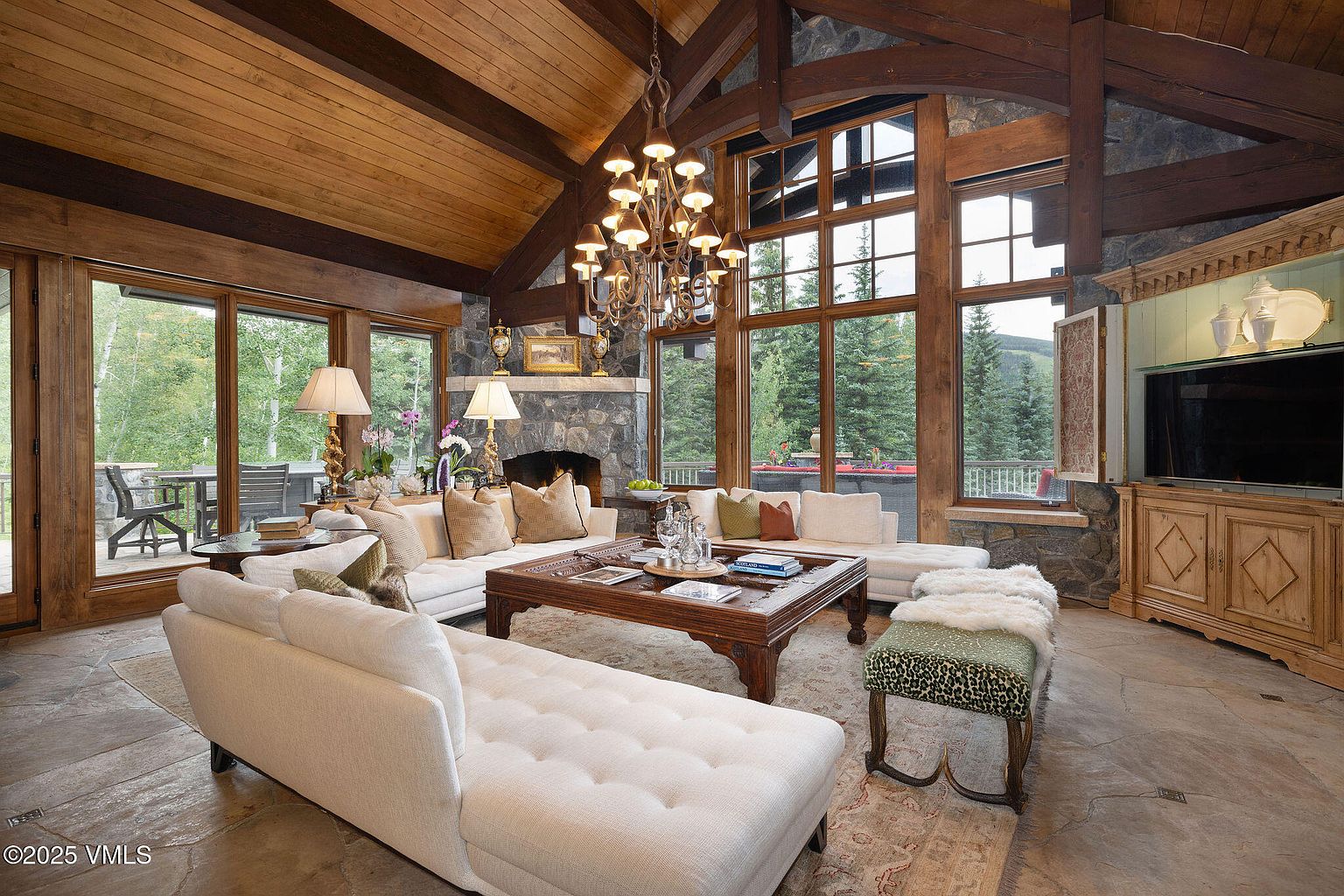 This is a spacious living room featuring a high, wood-paneled ceiling with exposed beams and a large stone fireplace. The room is furnished with comfortable white sofas, a large wooden coffee table, and a patterned ottoman. Large windows offer views of the surrounding trees, creating a cozy and luxurious atmosphere.