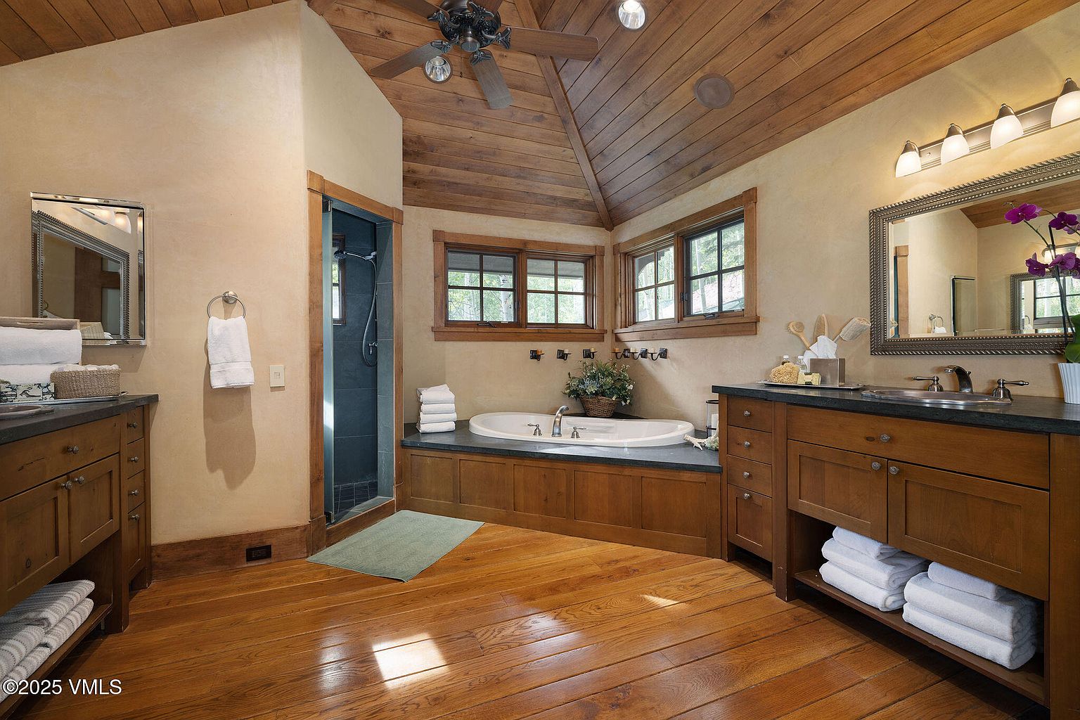 This is a primary bathroom featuring wood floors and a wood-paneled ceiling. The bathroom includes a bathtub with a dark countertop, a shower with a glass door, and a vanity with a mirror and lighting. The style is rustic and luxurious, creating a spa-like atmosphere.