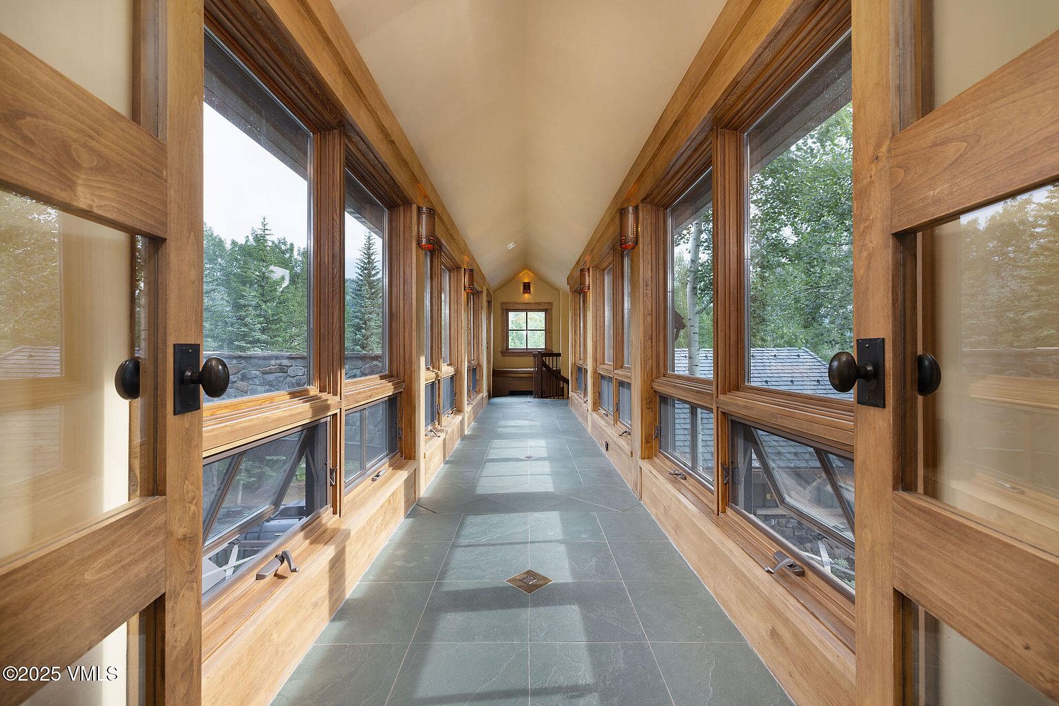 This interior shot showcases a long hallway with wooden-framed windows on both sides, offering views of the surrounding greenery. The hallway features a tiled floor and a light-colored ceiling, creating a bright and airy atmosphere. The open wooden doors at the entrance invite viewers into the space, highlighting the architectural details and natural light.