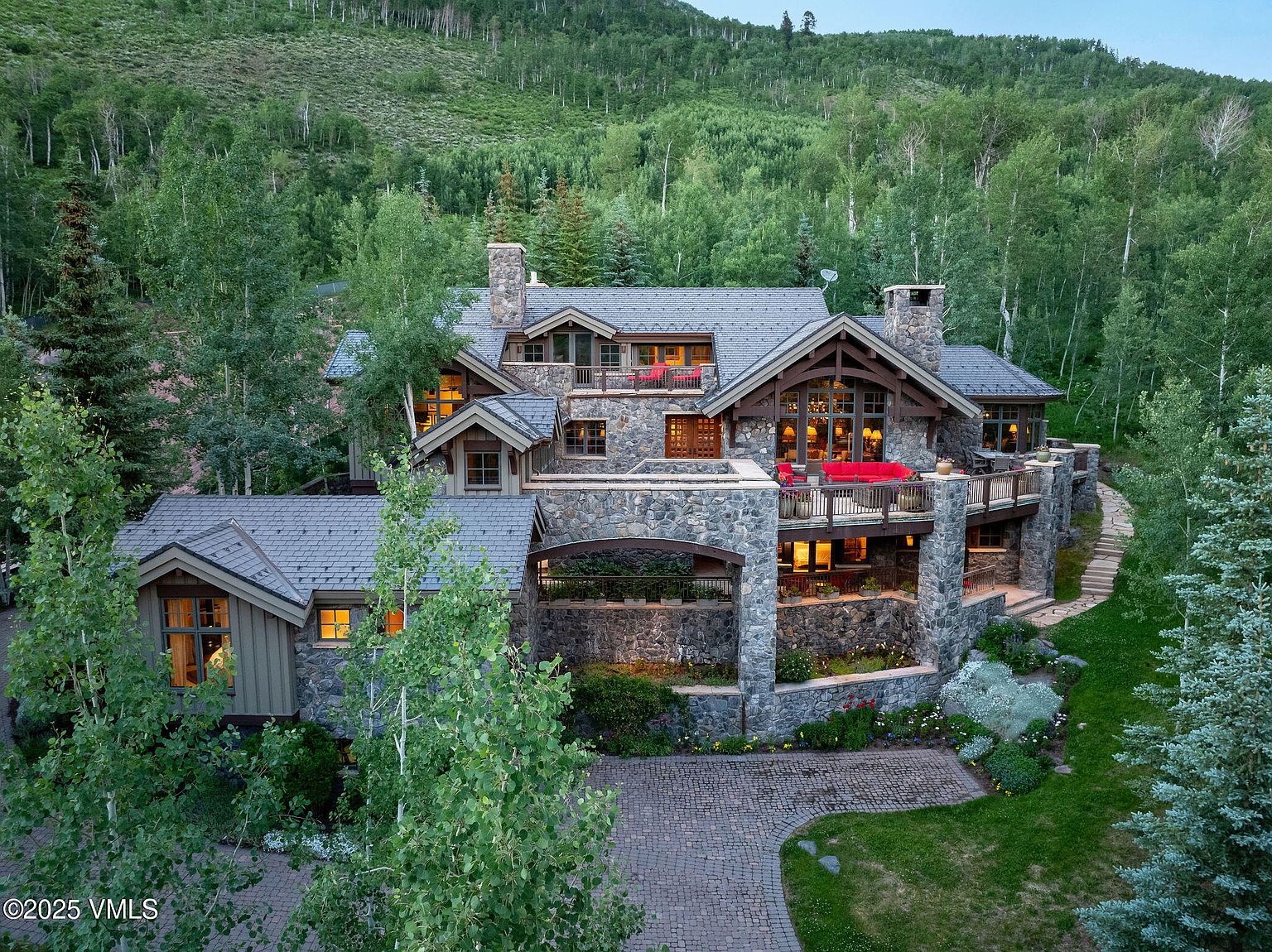 This aerial view showcases a grand stone and timber home nestled among lush green trees. The multi-level structure features multiple balconies with red outdoor furniture, a stone facade, and a meticulously landscaped yard with a brick driveway. The overall impression is one of luxury and seclusion in a natural setting.