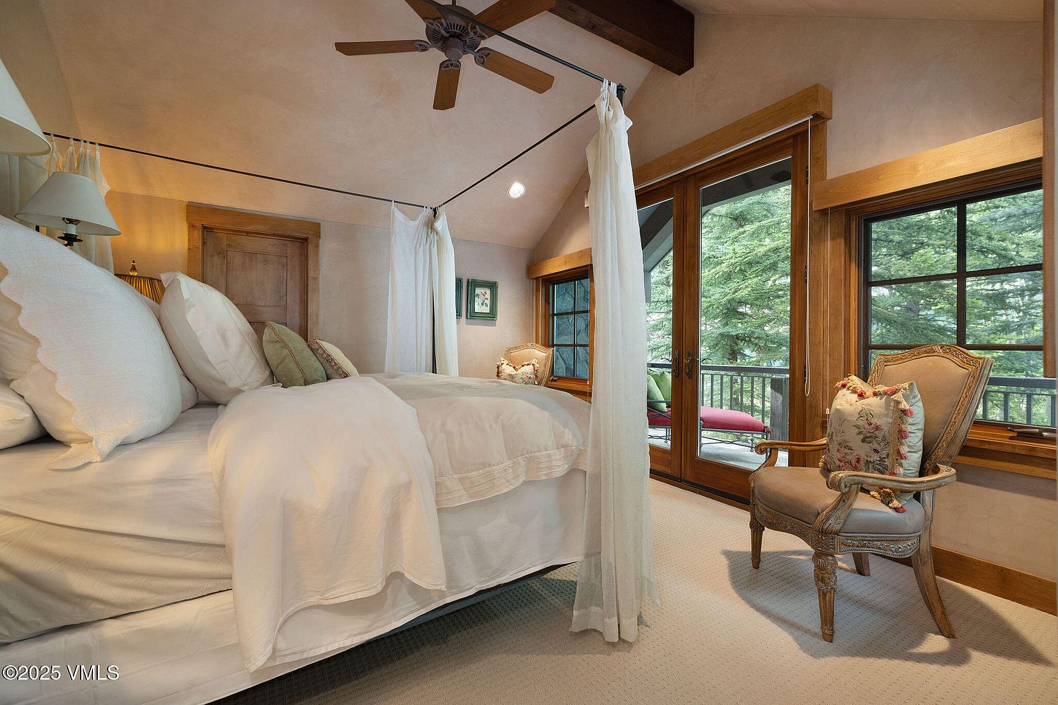 This is a primary bedroom featuring a four-poster bed with sheer white curtains, creating a soft and romantic ambiance. The room has a neutral color palette with beige walls and carpet, complemented by wooden window frames and a ceiling fan. A chair with a floral pillow sits near the window, offering a cozy reading spot with a view of the trees outside.
