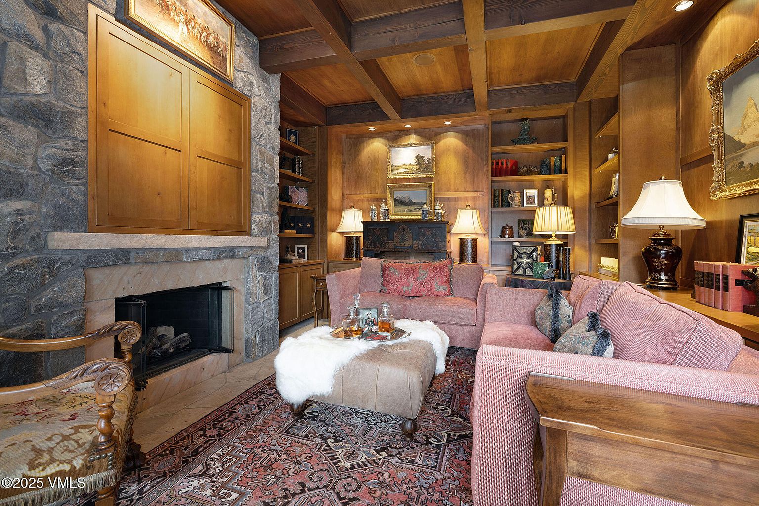 This is an interior shot of a cozy living room featuring a stone fireplace with a wooden cabinet above, flanked by built-in wooden bookshelves. The room is furnished with comfortable pink sofas, a leather ottoman with a fur throw, and a patterned rug, creating a warm and inviting atmosphere. The wooden beamed ceiling adds to the room's rustic charm.