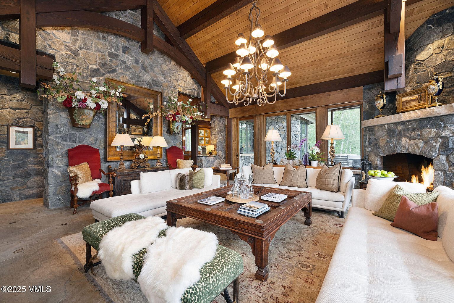 This is a spacious living room featuring a stone fireplace and walls, complemented by wooden beams and a ceiling. The room is furnished with comfortable white sofas, a large wooden coffee table, and decorative ottomans. A grand chandelier hangs from the ceiling, adding a touch of elegance to the rustic setting.