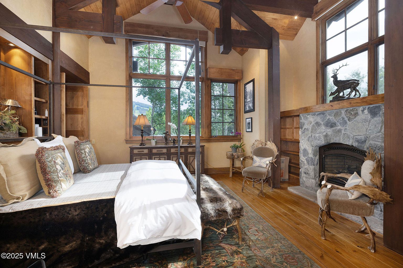 This is a luxurious primary bedroom featuring a four-poster bed with plush bedding and decorative pillows. The room boasts exposed wooden beams, a stone fireplace with a deer statue on the mantel, and large windows offering natural light. The overall style is rustic and elegant, creating a warm and inviting atmosphere.
