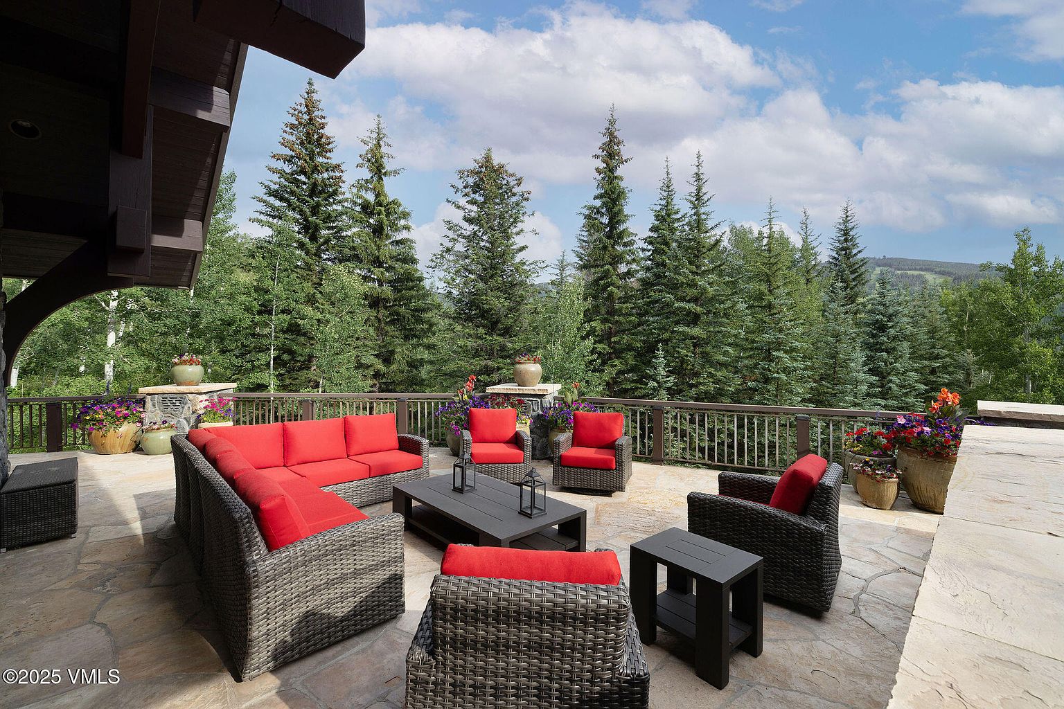 This image showcases a beautifully arranged outdoor patio area, featuring a wicker sectional sofa with red cushions, matching armchairs, and a dark-colored coffee table. The patio is surrounded by a wooden railing, offering views of lush green trees and a partly cloudy sky. The setting suggests a relaxing and inviting outdoor living space.
