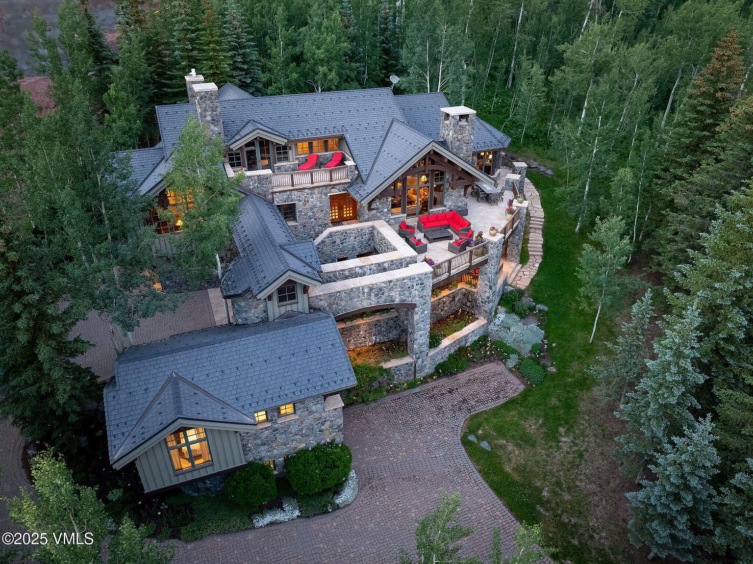 This aerial view showcases a luxurious stone-clad home nestled among lush trees. The property features multiple levels of outdoor living spaces, including balconies and patios with red furniture, a stone driveway, and a meticulously landscaped yard. The architecture blends seamlessly with the natural surroundings, creating a serene and private estate.