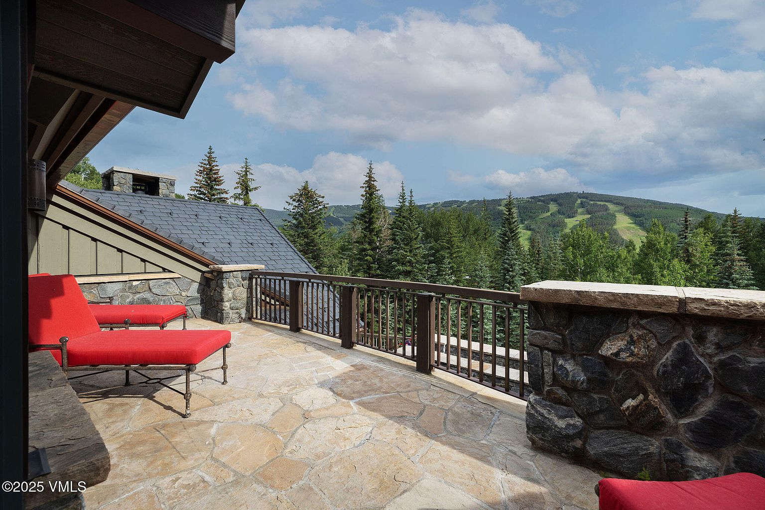 This image showcases a spacious patio or balcony with stone flooring and a rustic stone railing. Red lounge chairs provide seating, and the view overlooks a lush green mountain landscape with visible ski runs. The overall impression is one of luxury and relaxation, perfect for enjoying the natural surroundings.