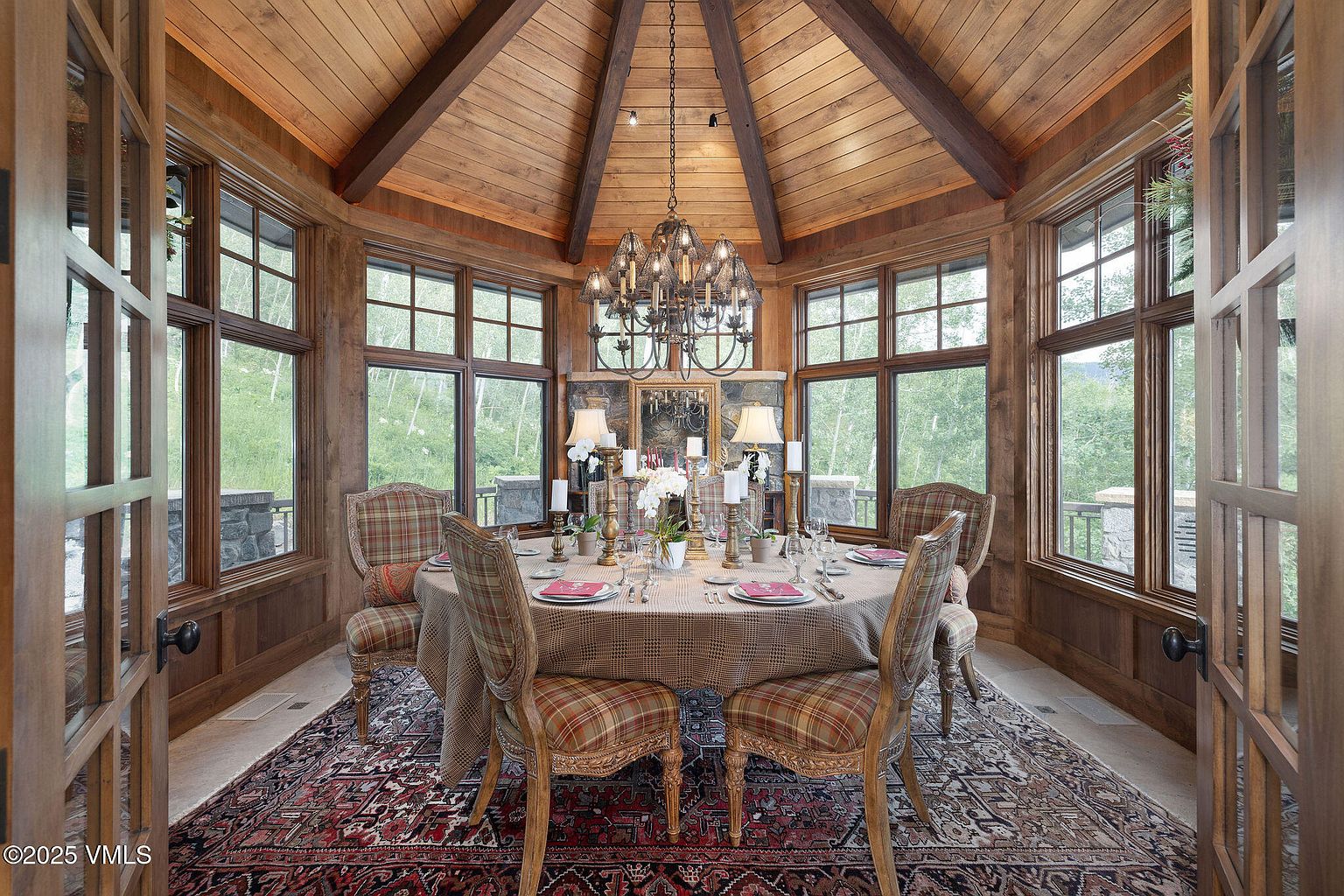 This is an elegant dining room featuring a round table set for a meal, surrounded by plaid chairs. The room is enclosed by large windows offering natural light and views of the surrounding landscape. The wood-paneled walls and ceiling, along with the ornate chandelier, contribute to a luxurious and inviting atmosphere.