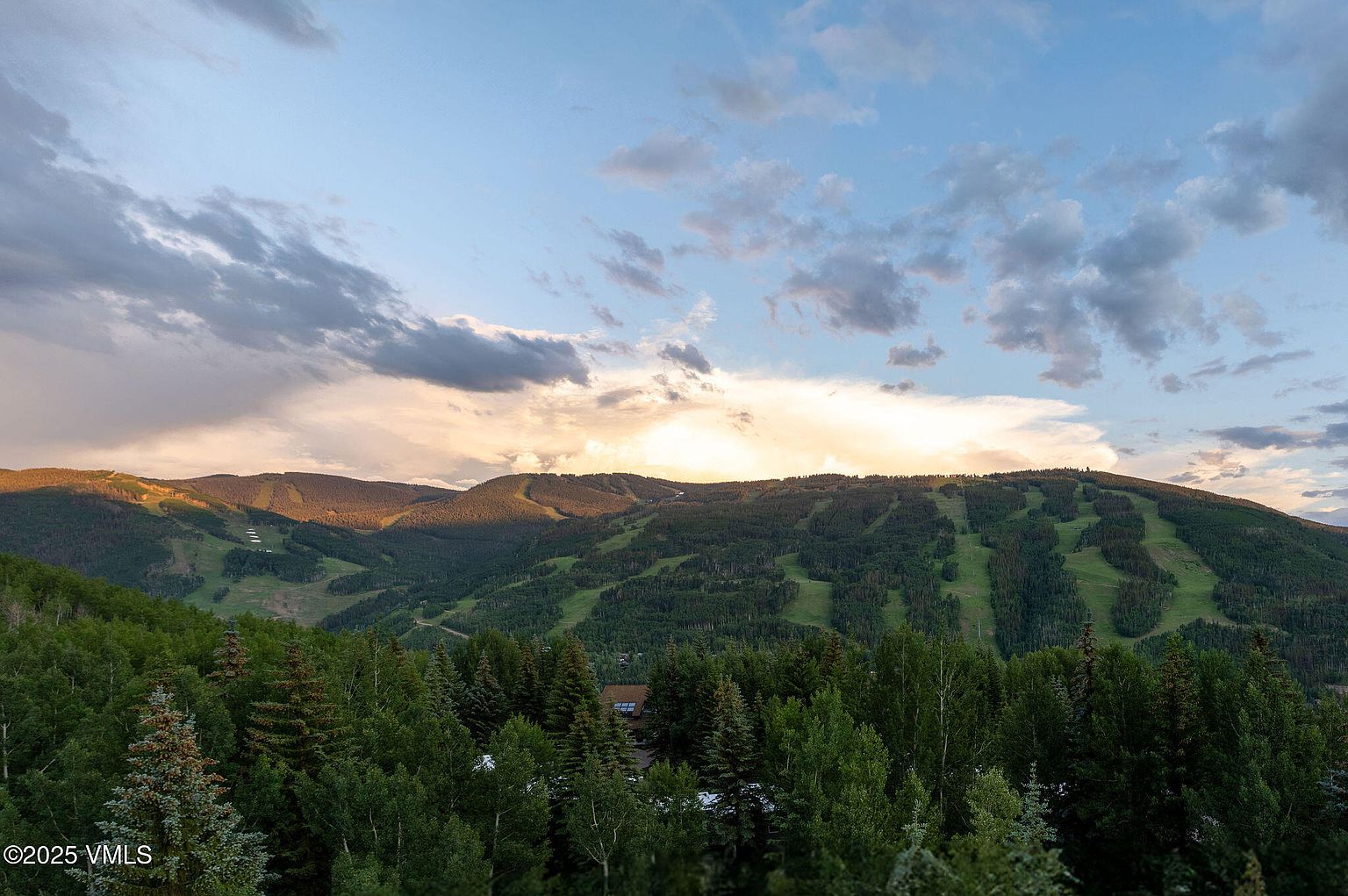 This aerial view showcases a stunning mountain landscape with lush green forests and ski slopes. The sky is filled with dramatic clouds, illuminated by the setting sun, creating a warm and inviting atmosphere. The image captures the natural beauty and recreational opportunities of the area, making it ideal for a real estate listing highlighting the location's scenic appeal.