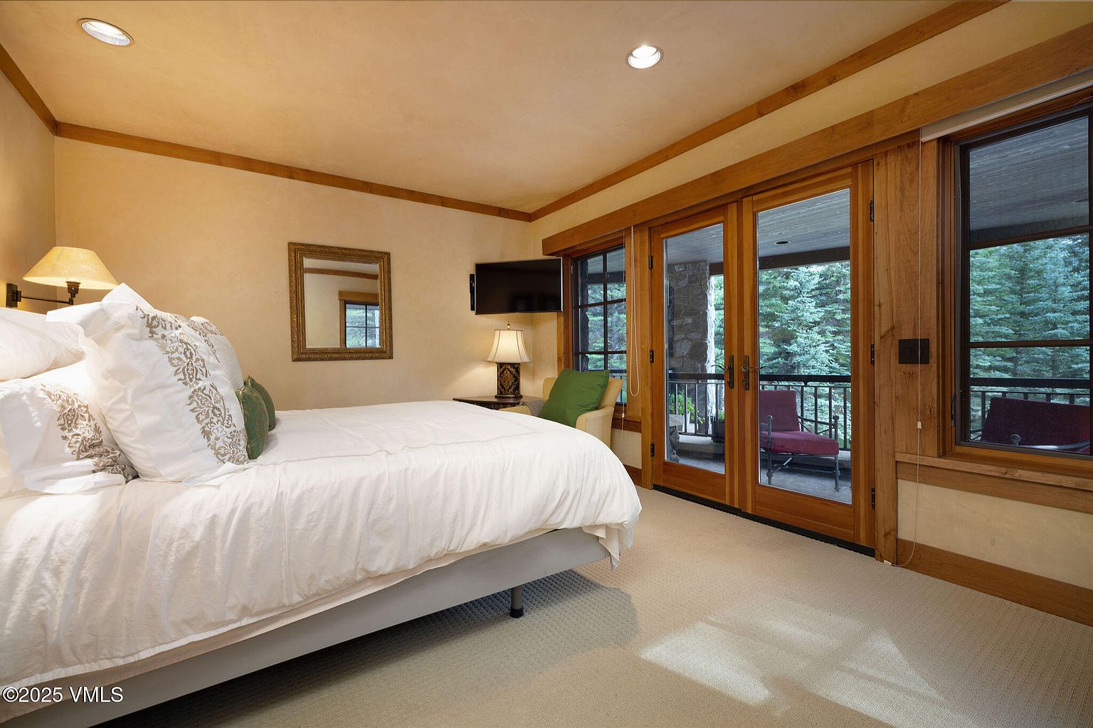 This is a primary bedroom featuring a king-size bed with white linens and decorative pillows. The room has wood trim, a mirror, and a flat-screen TV. Sliding glass doors lead to a balcony with views of the surrounding trees, creating a serene and inviting atmosphere.