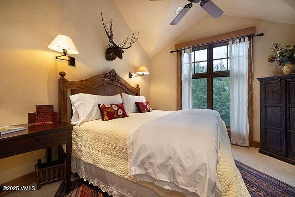This is a cozy primary bedroom featuring a wooden bed frame with decorative carvings, adorned with white linens and patterned pillows. A wooden desk sits to the side, and a mounted lamp provides warm lighting. The room has a rustic, inviting feel, enhanced by the mounted deer antlers and the view from the window.