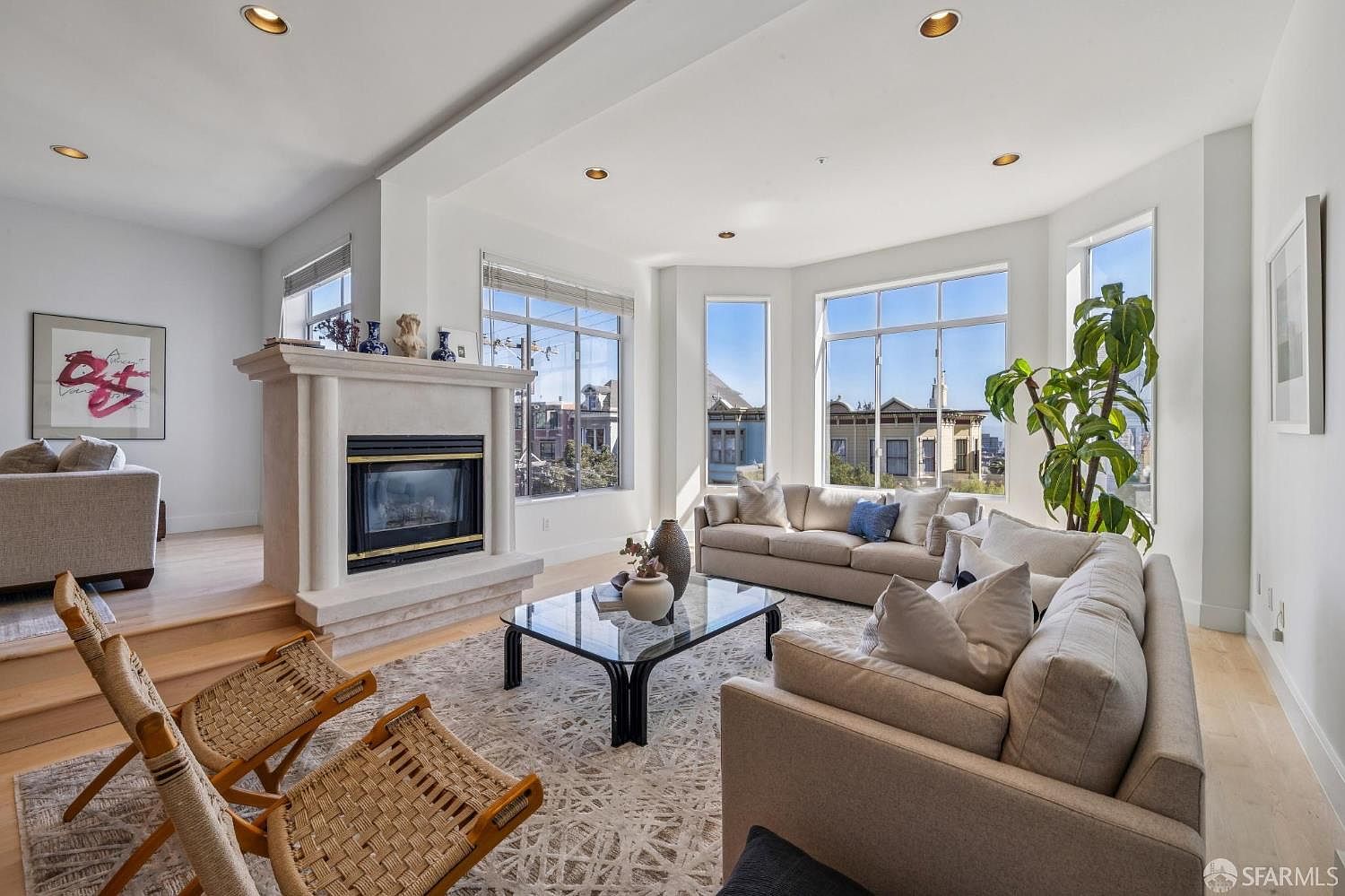This bright and airy living room features a contemporary design with a central fireplace acting as a focal point between two distinct seating areas. Large windows flood the space with natural light, offering views of the surrounding neighborhood, while a neutral color palette and textured rug create a sophisticated, inviting atmosphere. The open-concept layout seamlessly connects the spaces, highlighting the home's spacious and modern aesthetic.