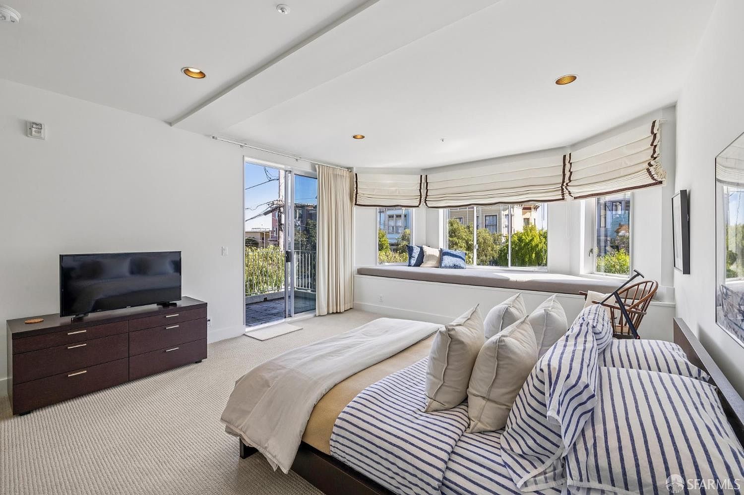 This bright and airy primary bedroom features a large bay window with a built-in bench seat, offering a cozy nook with natural light. The room is furnished with a bed dressed in blue and white striped linens, a dark wood dresser, and a television, all set against a neutral color palette. A sliding glass door leads out to a private balcony, enhancing the indoor-outdoor flow of the space.