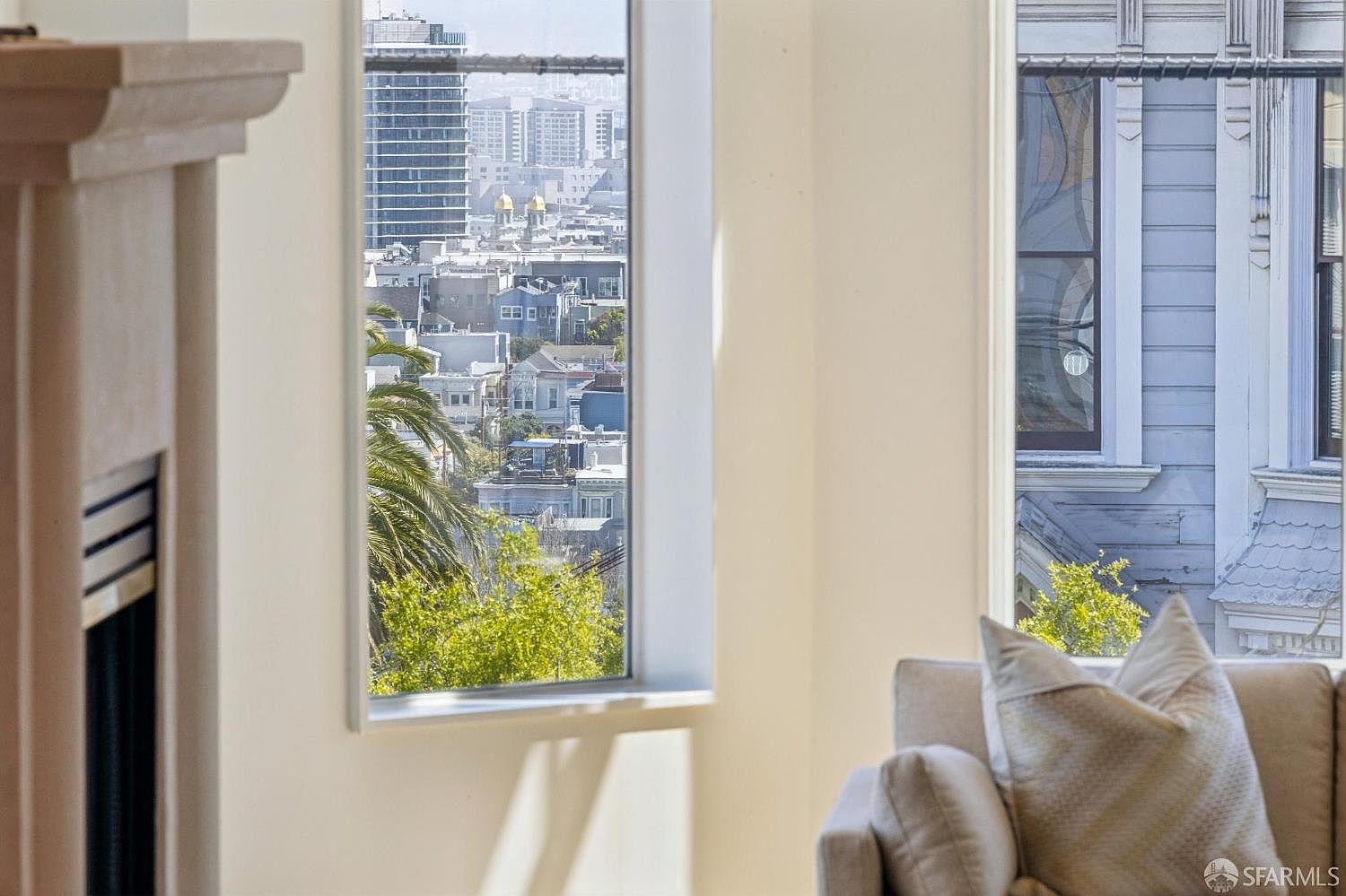 This interior shot captures a bright living room space, featuring a comfortable sofa with patterned pillows in the foreground. A large, floor-to-ceiling window serves as the focal point, framing a stunning urban cityscape view that includes high-rise buildings and residential rooftops. The clean, minimalist aesthetic of the room allows the natural light and the expansive city vista to take center stage.
