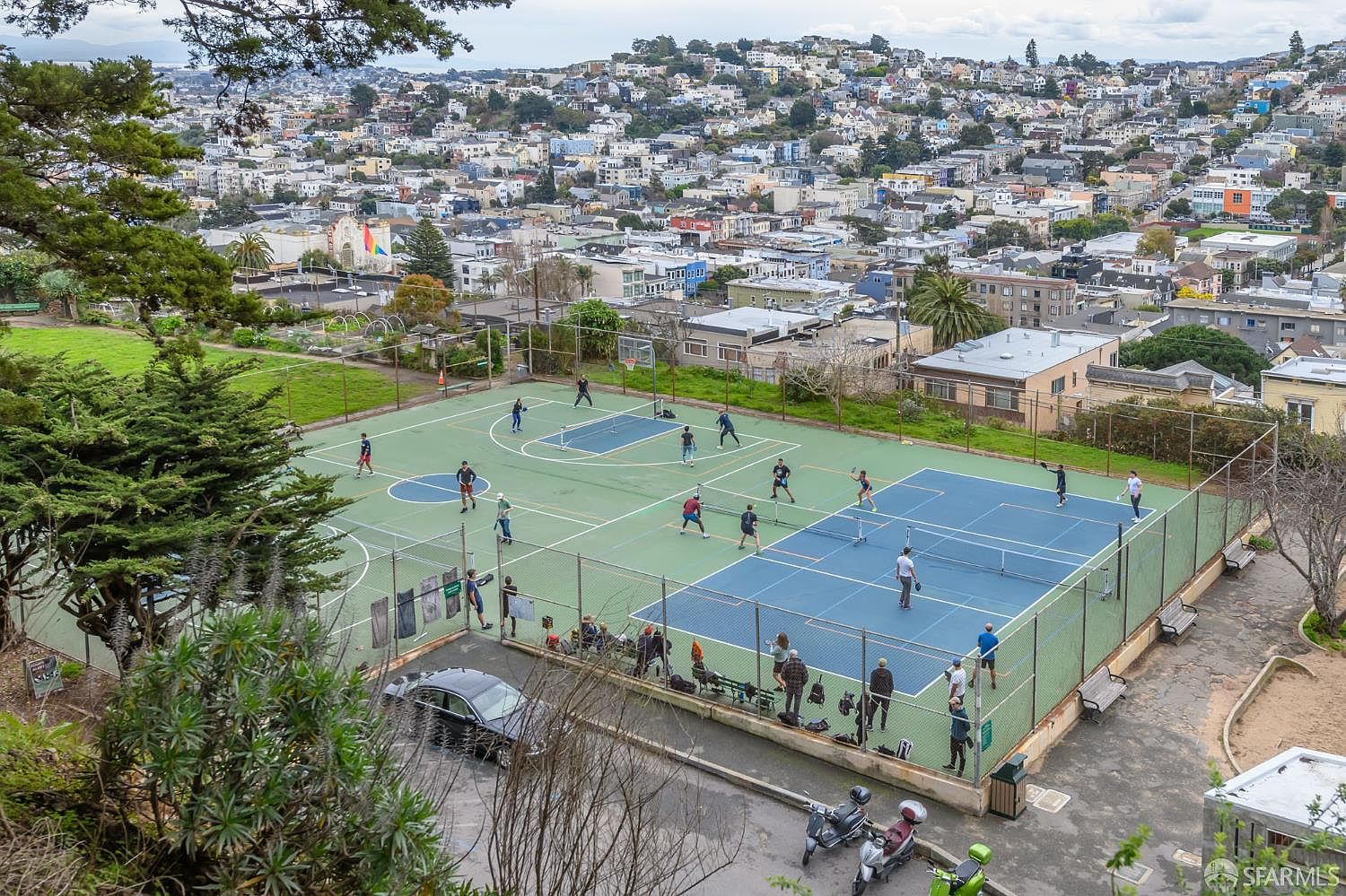 This high-angle aerial view captures a vibrant public park featuring multiple pickleball courts filled with active players. The scene is set against a dense urban backdrop of hillside homes, showcasing the community's recreational lifestyle. The perspective provides a clear overview of the facility's layout, fencing, and surrounding neighborhood context.