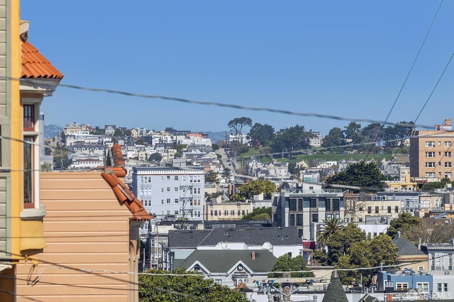 This elevated, wide-angle shot captures a dense urban cityscape, likely in San Francisco, characterized by a mix of historic Victorian architecture and modern apartment buildings. The perspective looks out from a high vantage point, showcasing the rolling hills, layered rooftops, and distant greenery under a clear blue sky. The foreground features the corner of a tan-colored building with a terracotta roof, framing the expansive view of the neighborhood.