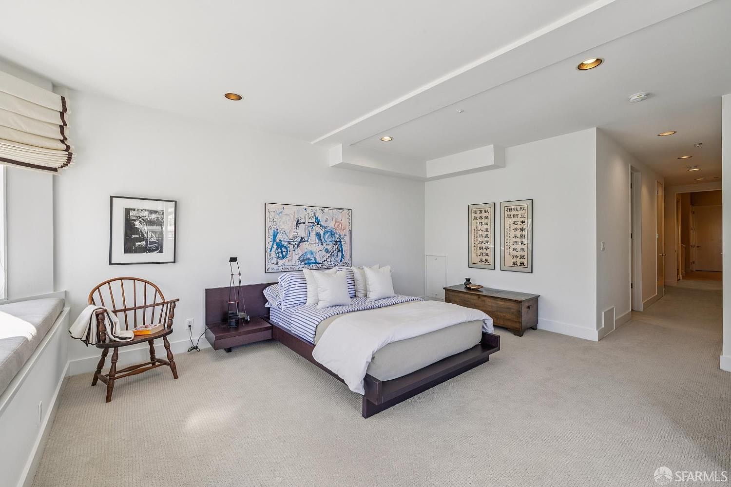 This spacious primary bedroom features a modern platform bed with striped bedding, flanked by a dark wood nightstand and a matching wooden storage chest. The room is decorated with minimalist art, including two framed calligraphic pieces and a large abstract painting, all set against crisp white walls and neutral carpeting. A classic wooden spindle chair sits near a window nook, creating a serene and sophisticated atmosphere.