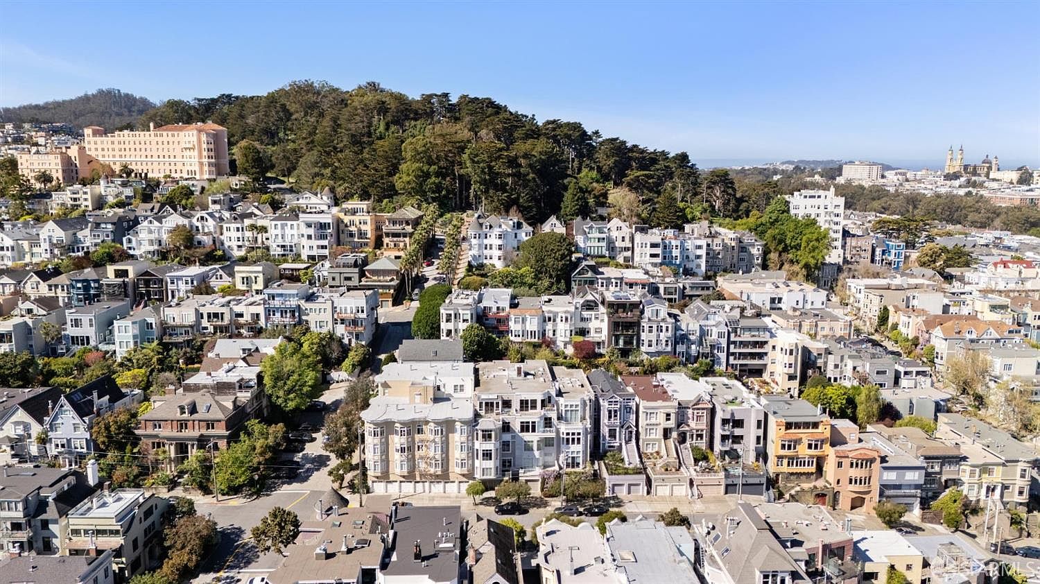 This high-angle aerial view captures a dense, historic urban neighborhood in San Francisco, characterized by a mix of Victorian and Edwardian-style architecture. The scene showcases rolling hills covered in lush greenery, with iconic city landmarks visible in the distance under a clear blue sky. The perspective emphasizes the tight-knit layout of the residential streets and the unique, layered topography of the city.