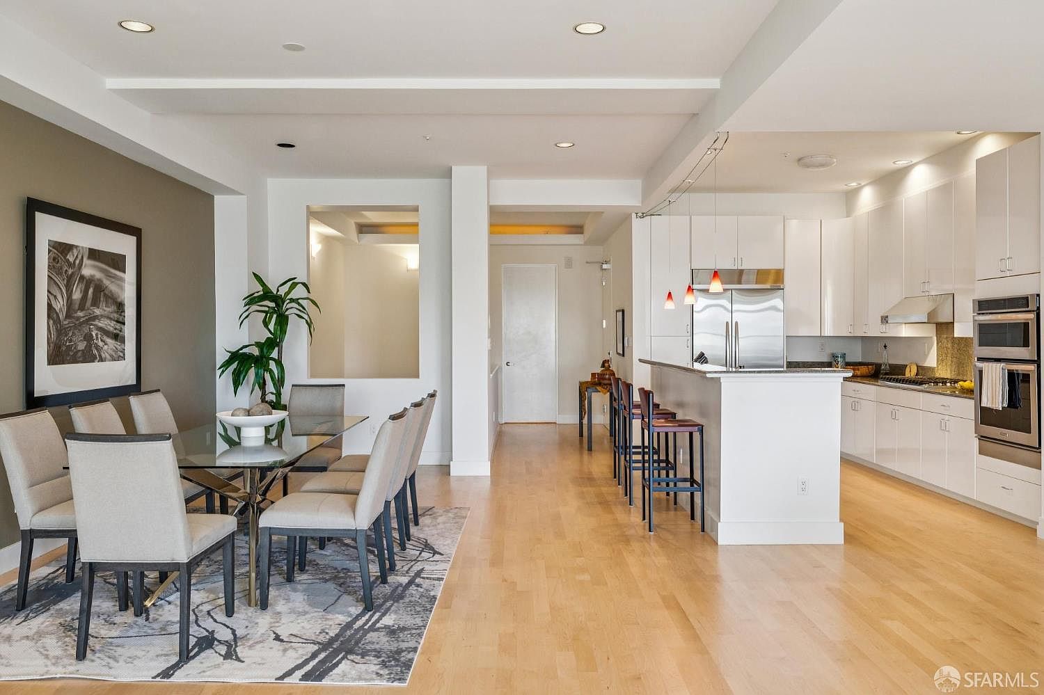 This open-concept space seamlessly blends a modern dining area with a sleek, contemporary kitchen. The dining room features a glass-topped table surrounded by upholstered chairs on a patterned area rug, while the adjacent kitchen boasts white cabinetry, stainless steel appliances, and a breakfast bar with stools. The perspective is a wide-angle shot from the dining area looking toward the kitchen, emphasizing the airy, light-filled layout and hardwood flooring.