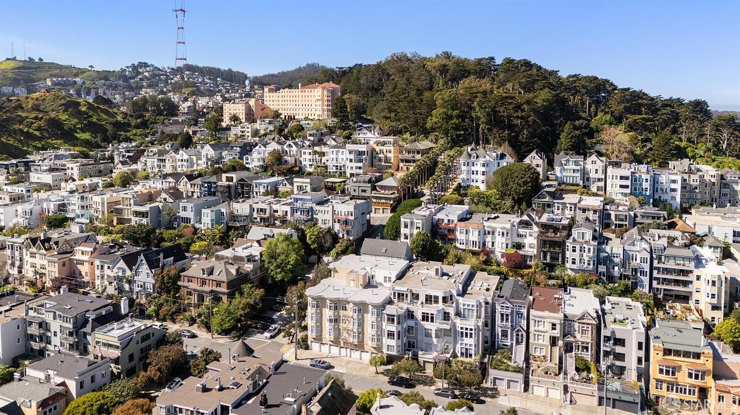 This high-angle aerial view captures a dense, picturesque neighborhood in San Francisco, characterized by its iconic Victorian and Edwardian architecture nestled against a lush, tree-covered hillside. The scene showcases a complex urban landscape with winding streets, multi-story residential buildings, and a prominent radio tower in the distance. The bright, clear daylight emphasizes the architectural variety and the hilly topography typical of the city's unique residential districts.