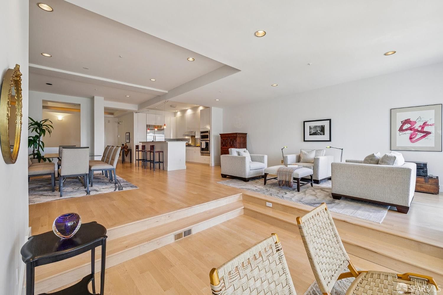 This spacious, open-concept living area features a sunken living room design with light hardwood flooring and neutral-toned furniture. The space seamlessly transitions into a dining area and a modern kitchen, creating an airy and sophisticated atmosphere perfect for entertaining. The perspective is from a slightly elevated vantage point, looking down into the living room, which highlights the architectural depth and clean, contemporary aesthetic of the home.
