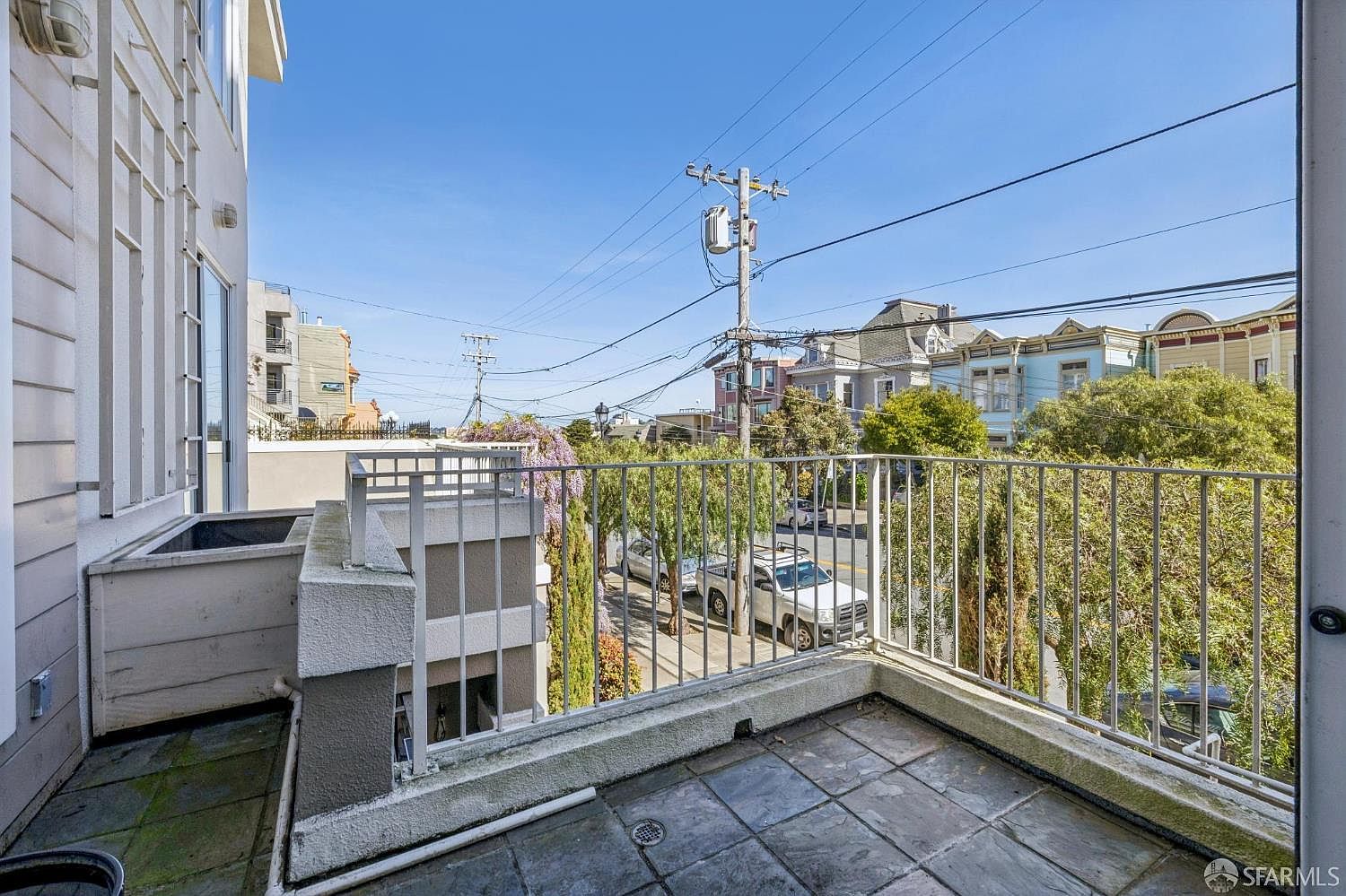 This image captures a private balcony with tiled flooring and a metal railing, offering an elevated view of a quiet residential street. The scene features a clear blue sky, mature trees, and neighboring historic-style homes, creating a bright and airy atmosphere. The perspective is from the balcony looking outward, emphasizing the outdoor living space and the surrounding neighborhood character.