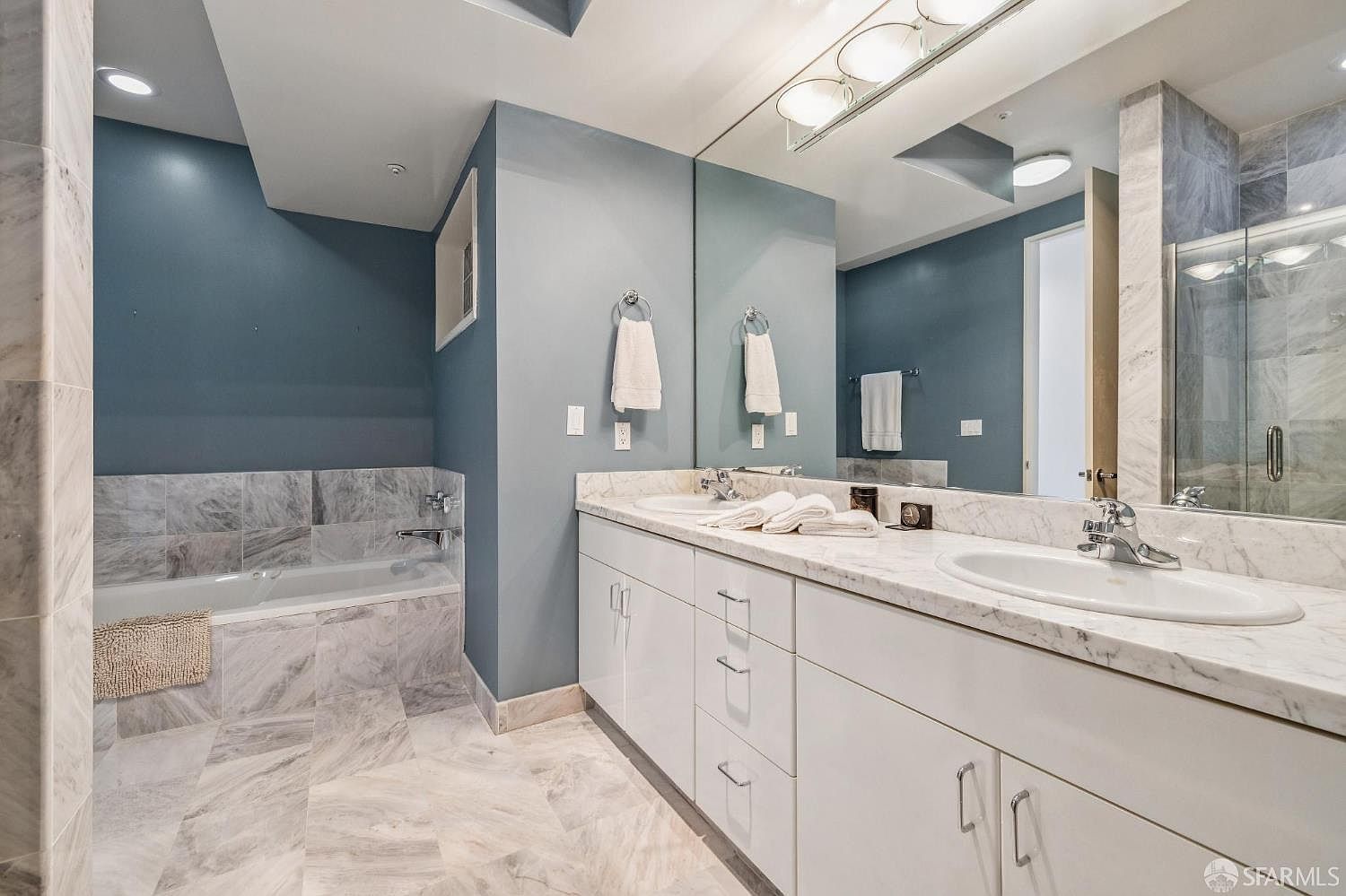 This primary bathroom features a clean, contemporary design with a dual-sink vanity topped with white marble countertops and white cabinetry. A large soaking tub is encased in matching marble tile, complemented by blue-toned walls and a spacious wall-to-wall mirror. The room is well-lit by overhead vanity lighting and recessed ceiling lights, creating a bright and polished atmosphere.