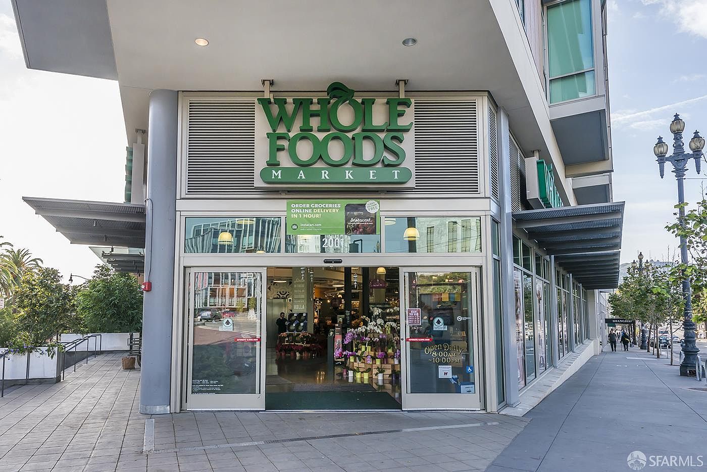 This image captures the front entrance of a Whole Foods Market, showcasing a modern commercial storefront with large glass doors and windows. The building features a clean, contemporary architectural style with a prominent green logo sign above the entrance and a paved sidewalk leading up to the doors. The perspective is a straight-on, eye-level shot that highlights the accessibility and inviting nature of the retail space.