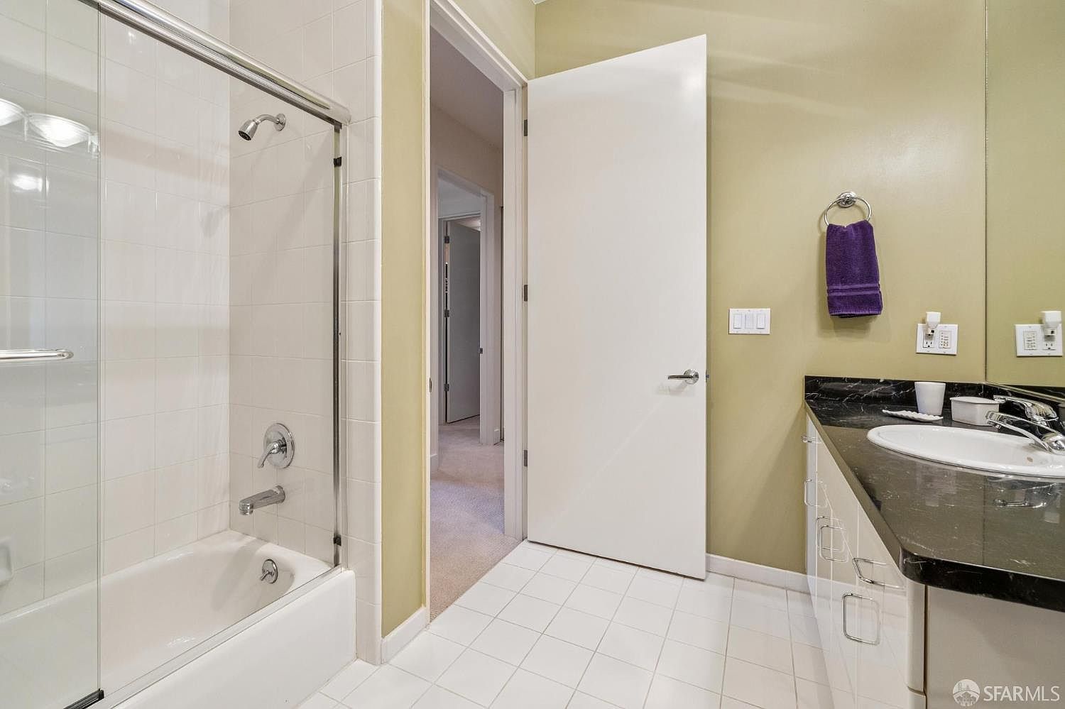This clean and functional bathroom features a shower-tub combination with a glass enclosure and white tile surround. A dark granite countertop with a white undermount sink provides a sharp contrast against the light-colored walls and white floor tiles. The space is well-lit and includes a purple hand towel, creating a simple and tidy aesthetic.