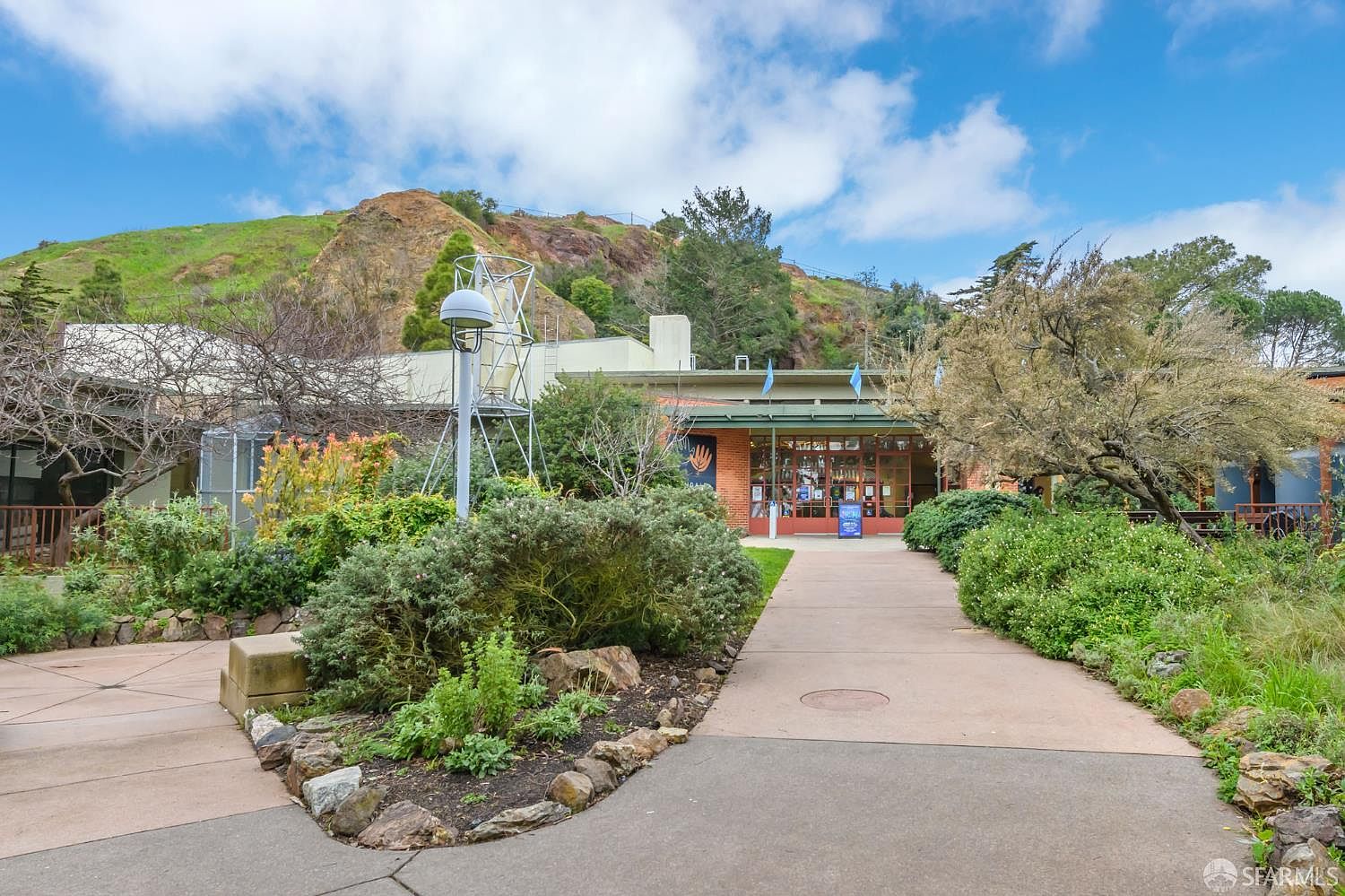 This image captures the inviting entrance of a community center or park facility, nestled against a scenic, grassy hillside. A paved walkway leads directly to the building's glass-fronted entrance, flanked by lush, mature landscaping and native shrubs. The scene is bathed in bright, natural daylight, creating a welcoming and serene atmosphere perfect for a public or community space.