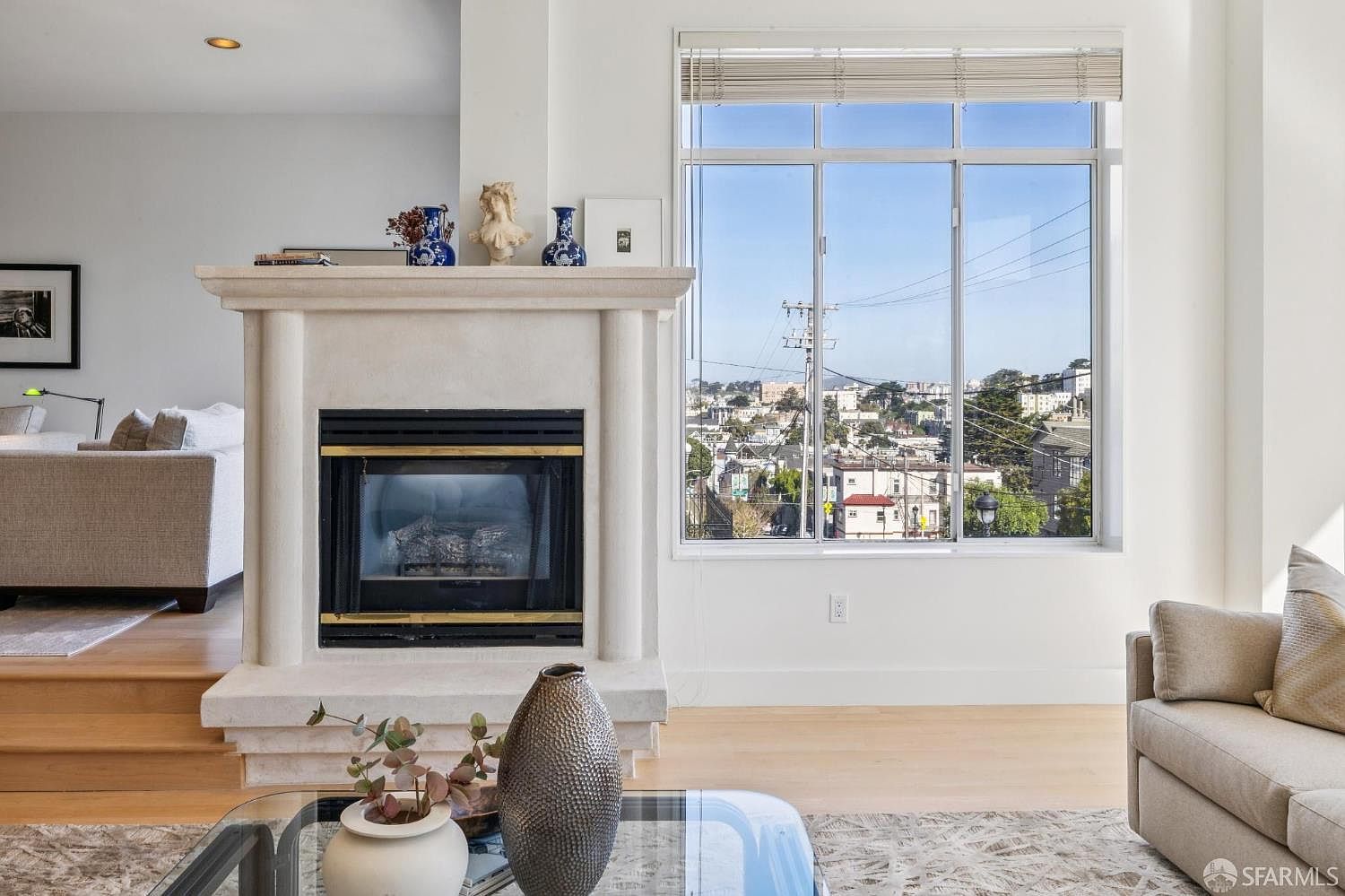 This bright and airy living room features a prominent central fireplace with a classic mantle, serving as a focal point between the seating area and a large window. The room offers an expansive view of the city skyline, creating a sophisticated urban atmosphere. The neutral color palette and clean lines contribute to a modern, elegant aesthetic suitable for high-end living.