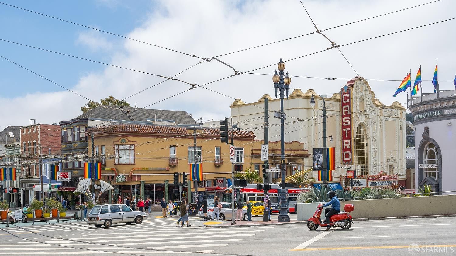This image captures a vibrant street-level view of the iconic Castro Theatre and surrounding neighborhood in San Francisco. The scene features a bustling intersection with a red scooter in the foreground, classic architecture, and colorful pride flags adorning the streetscape. The perspective is a wide-angle shot that emphasizes the lively urban atmosphere and historic character of this famous community landmark.