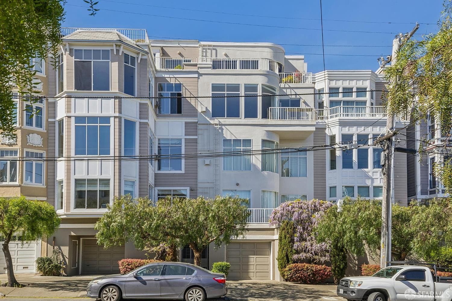 This image captures the front facade of a multi-story residential building, showcasing a contemporary architectural style with a mix of beige siding and stucco finishes. The structure features prominent bay windows, multiple balconies, and integrated garage doors at the street level, framed by mature trees and flowering bushes. The perspective is a straight-on, eye-level shot that highlights the building's symmetrical design and urban curb appeal.