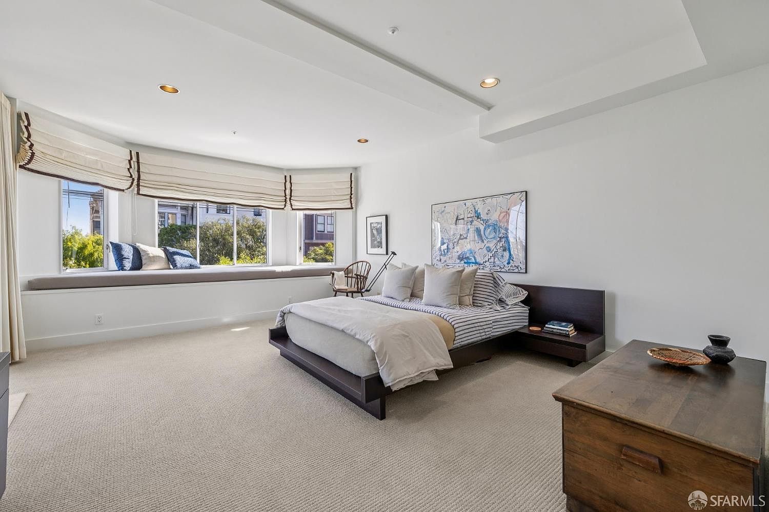 This spacious primary bedroom features a modern platform bed with a dark wood frame, centered against a minimalist wall adorned with abstract art. A large, curved window seat with plush cushions provides a cozy reading nook overlooking the outdoors, while neutral carpeting and recessed lighting create a serene, airy atmosphere. The room is accented by a rustic wooden chest in the foreground, adding a touch of warmth to the clean, contemporary design.