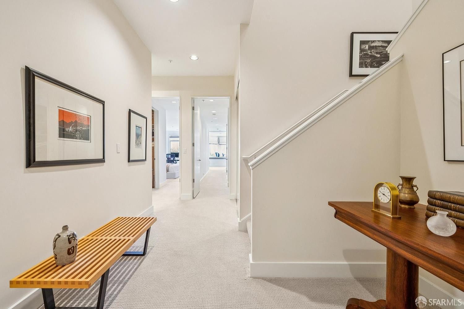 This bright, neutral-toned hallway features a clean, modern aesthetic with light carpeting and white walls. A slatted wooden bench sits on the left, while a wooden console table with decorative accents anchors the right side near the staircase. The perspective looks down the corridor toward an open doorway, creating a sense of depth and flow throughout the home.