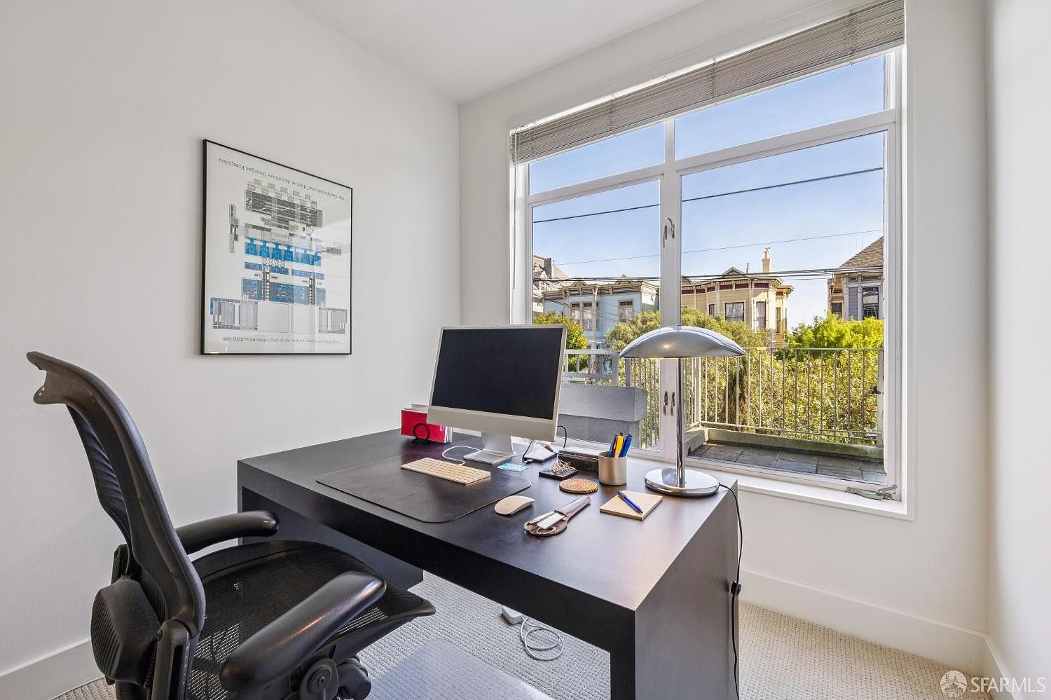 This bright and modern home office features a sleek black desk positioned to face a large window, which provides ample natural light and a view of the neighborhood. The room is minimalist in design, accented by a framed architectural print on the wall and a comfortable ergonomic office chair. The overall atmosphere is professional, quiet, and conducive to focused work.