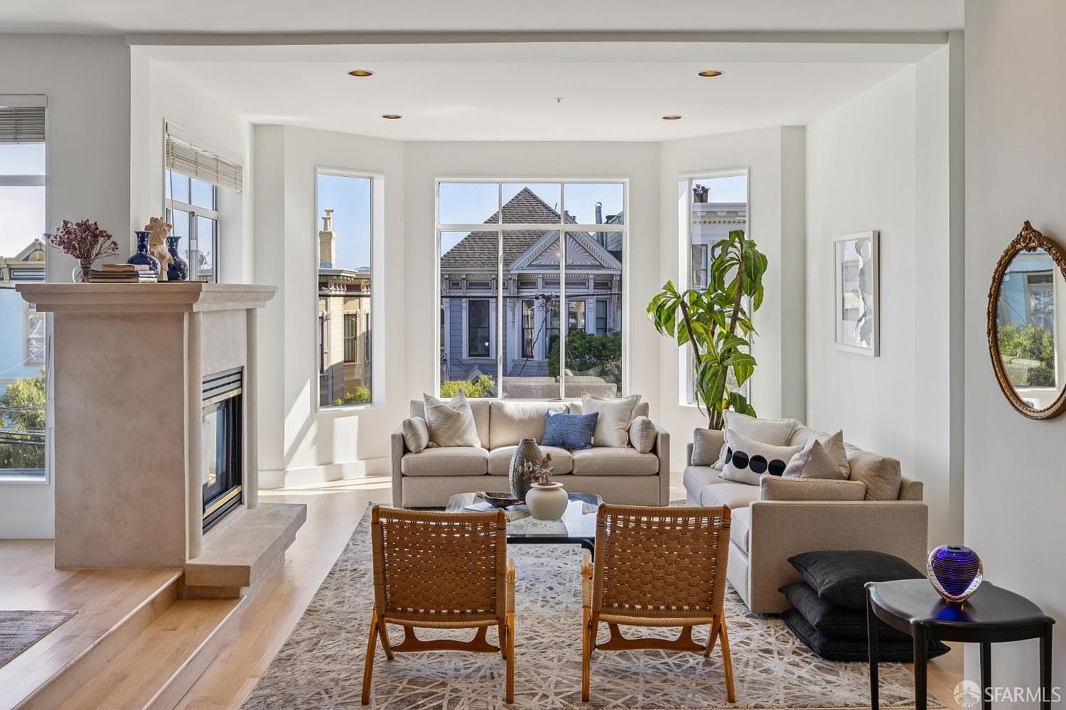 This bright and airy living room features a sophisticated, neutral color palette with plush seating arranged around a glass-topped coffee table. A prominent stone fireplace serves as a focal point on the left, while large bay windows flood the space with natural light and offer a charming view of the neighborhood architecture. The room is accented by a large indoor plant, a decorative mirror, and a textured area rug, creating a welcoming and elegant atmosphere.