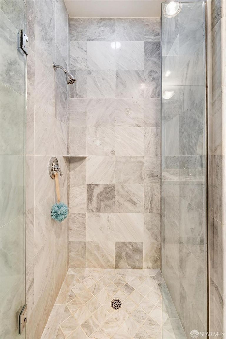 This image showcases a clean, modern walk-in shower featuring elegant, light-toned marble tile work on both the walls and the floor. The shower includes a glass door, a polished chrome showerhead, and a built-in corner shelf, creating a bright and spa-like atmosphere. The perspective is a straight-on shot looking into the shower enclosure, highlighting the high-quality materials and finishes.