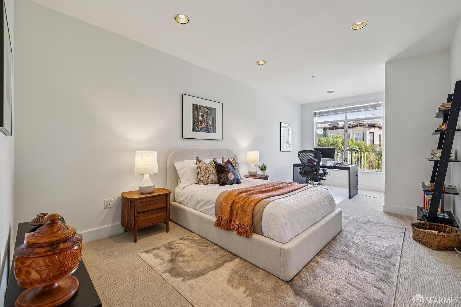 This bright and modern bedroom features a neutral color palette with a plush upholstered bed as the centerpiece, accented by a warm orange throw blanket. The room seamlessly integrates a functional home office area near the large window, which provides ample natural light and a view of the neighborhood. A sleek black ladder shelf and a decorative wooden urn add character to the space, creating a balanced and inviting atmosphere.