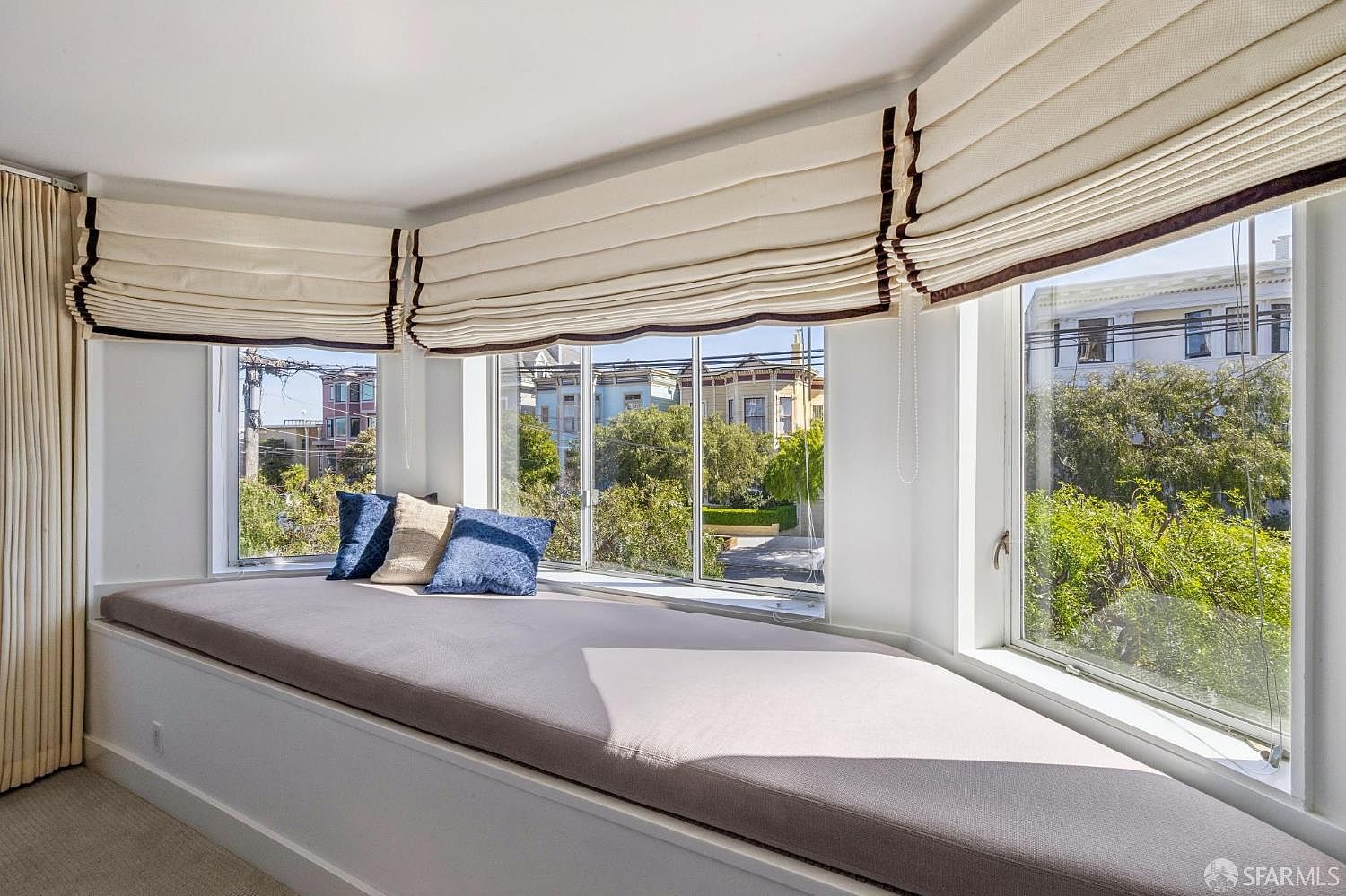 This image showcases a cozy, built-in window seat nestled within a bay window, offering a bright and inviting space for relaxation. The area is furnished with a plush, neutral-toned cushion and accented with decorative throw pillows, framed by elegant, cream-colored Roman shades. The perspective looks out through the large windows, providing a clear view of the surrounding neighborhood and lush greenery outside.