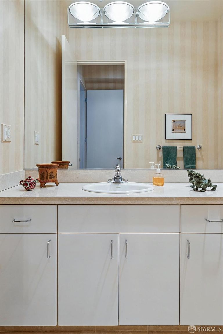 This bathroom features a clean, functional vanity with a white countertop, a single sink, and white cabinetry with silver hardware. The space is accented by striped wallpaper, a large wall-to-wall mirror, and a three-light vanity fixture above. Decorative items, including small figurines and a soap dispenser, add a personalized touch to the neutral, well-lit space.