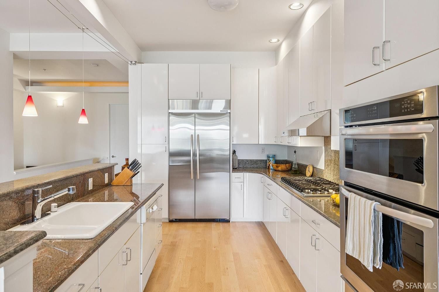 This modern, galley-style kitchen features sleek white cabinetry paired with dark granite countertops and light hardwood flooring. The space is equipped with high-end stainless steel appliances, including a double wall oven and a large refrigerator, while two red pendant lights add a pop of color over the breakfast bar. The perspective is a straight-on shot from the kitchen entrance, highlighting the clean lines and functional layout of the workspace.
