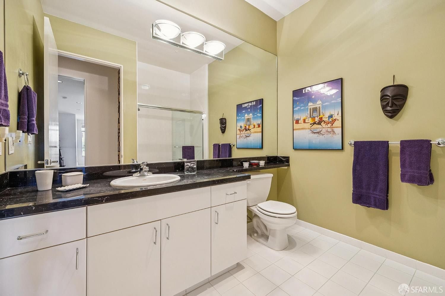This bathroom features a clean, functional design with a white vanity topped by a dark, speckled stone countertop and a large wall-to-wall mirror. The room is accented with sage green walls, purple towels, and decorative wall art, creating a personalized aesthetic. The space includes a standard toilet and a shower-tub combination, providing a practical layout for guests or secondary use.