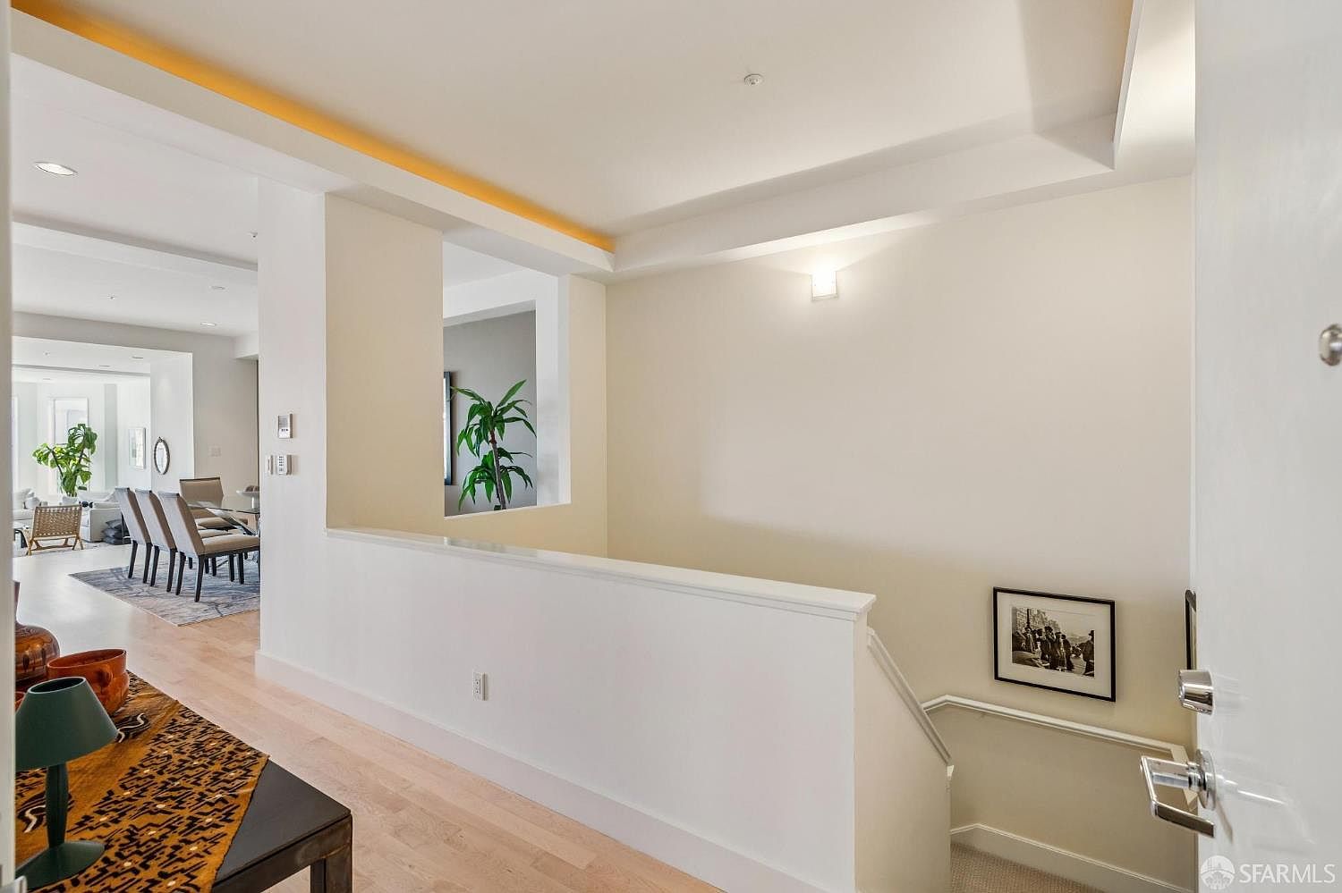 This interior view captures a bright, modern landing area that overlooks a staircase leading to a lower level. The space features clean white walls, light hardwood flooring, and a decorative cutout that provides a glimpse into the adjacent living area. The perspective is from the top of the stairs, emphasizing the open, airy flow of the home's floor plan.