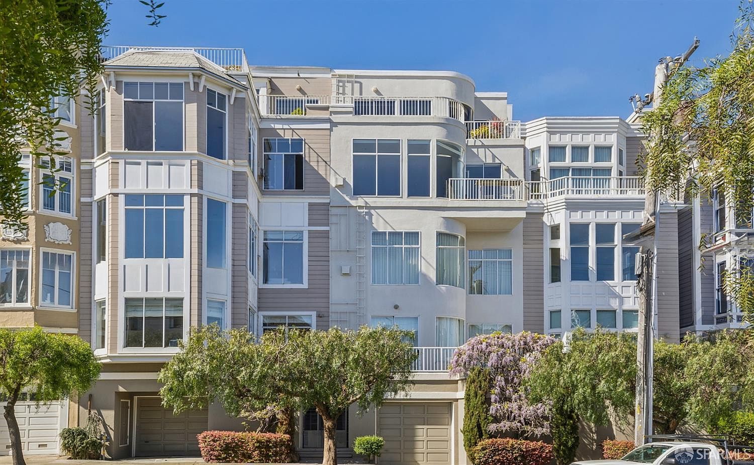 This image captures the front facade of a multi-story residential building, showcasing a blend of modern and traditional architectural elements with large windows and balconies. The structure features a light-colored exterior with distinct bay window sections and integrated garage doors at the street level. Lush green trees and flowering bushes in the foreground add a touch of natural beauty to the urban streetscape.