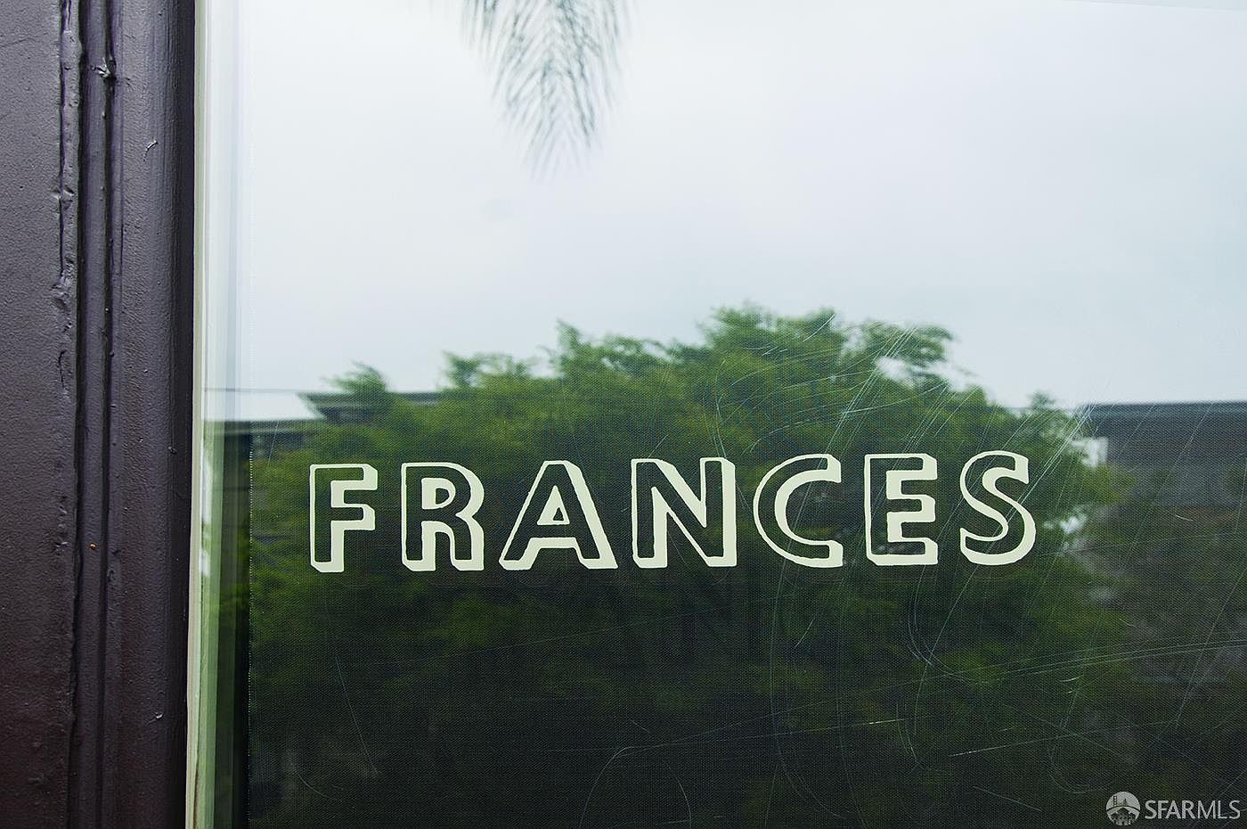 This image captures a close-up view of a glass door or window featuring the word 'FRANCES' in clean, white lettering. Through the glass, a lush green tree and a glimpse of a building structure are visible, creating a soft, natural backdrop. The perspective is intimate and focused, suggesting a welcoming entrance to a boutique or residential space.