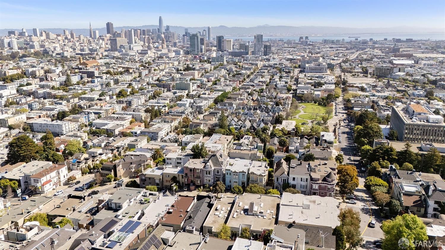 This high-altitude aerial shot captures a sprawling urban landscape, showcasing a dense neighborhood of residential buildings leading toward a prominent city skyline. The perspective emphasizes the scale of the city, highlighting the proximity of local parks and the architectural diversity of the surrounding homes. It provides a comprehensive overview of the neighborhood's layout and its integration into the broader metropolitan area.
