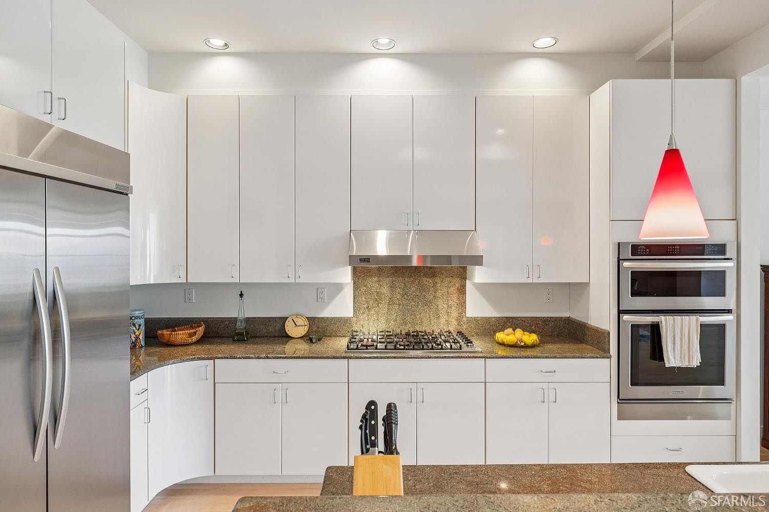 This modern kitchen features sleek white high-gloss cabinetry paired with dark granite countertops and a matching backsplash. The space is equipped with stainless steel appliances, including a built-in double oven and a large refrigerator, while a vibrant red pendant light adds a pop of color over the oven area. The perspective is a straight-on, eye-level shot that highlights the clean, contemporary design and functional layout of the culinary space.